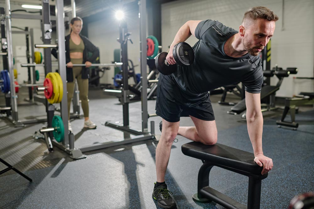 Man performing a one arm dumbbell row on a bench in a gym, with a woman lifting in the background.